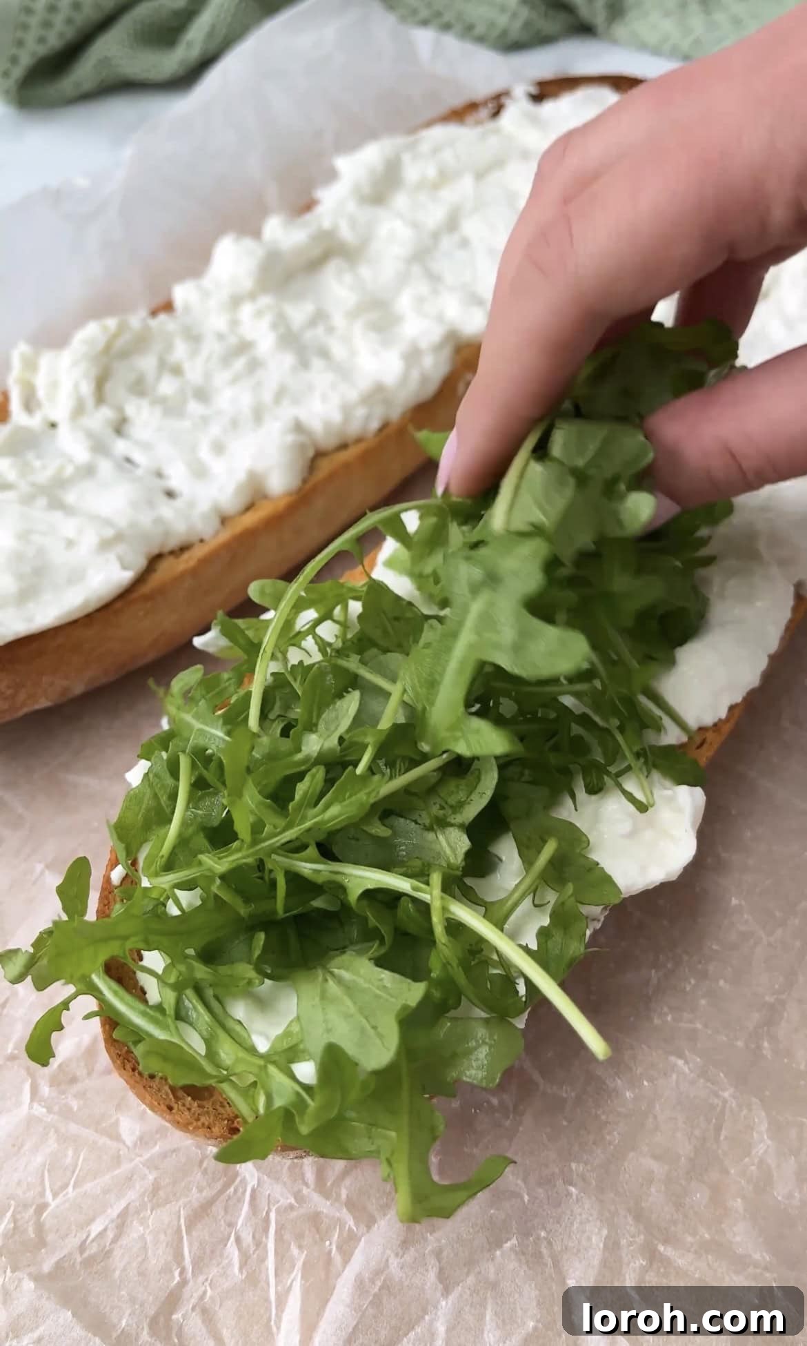 adding rocket. Fresh green rocket leaves being scattered generously over the burrata-topped ciabatta, adding a peppery layer.