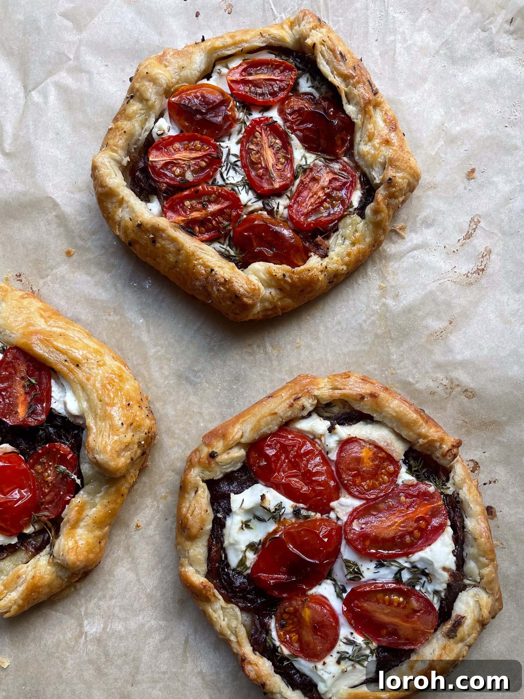 Sweet Onion and Goat Cheese Galettes with Sun-Kissed Cherry Tomatoes 4 Three perfectly baked individual galettes, generously topped with cherry tomatoes, goat cheese, and red onions, arranged neatly on baking parchment.