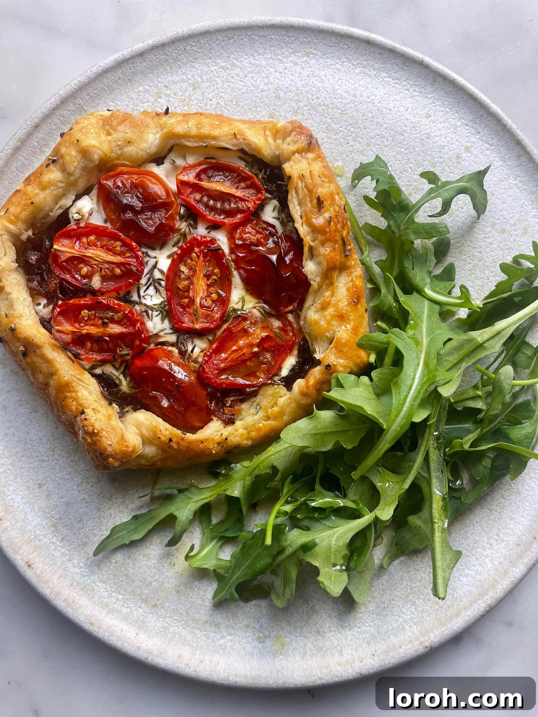 Sweet Onion and Goat Cheese Galettes with Sun-Kissed Cherry Tomatoes 5 A beautifully presented red onion, goat cheese, and cherry tomato galette resting on a grey plate, accompanied by a fresh side salad of rocket.