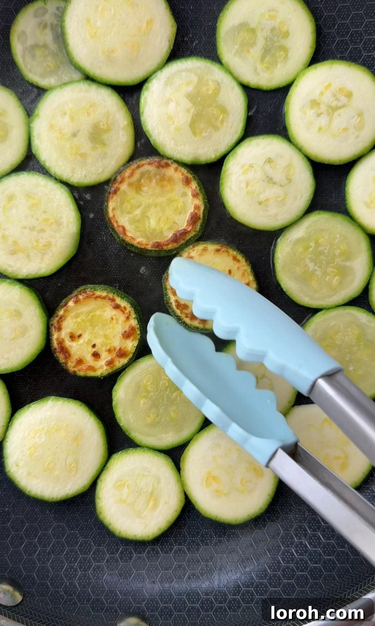 Slices of courgettes (zucchini) being gently fried until lightly golden and softened in a large pan with a drizzle of olive oil.
