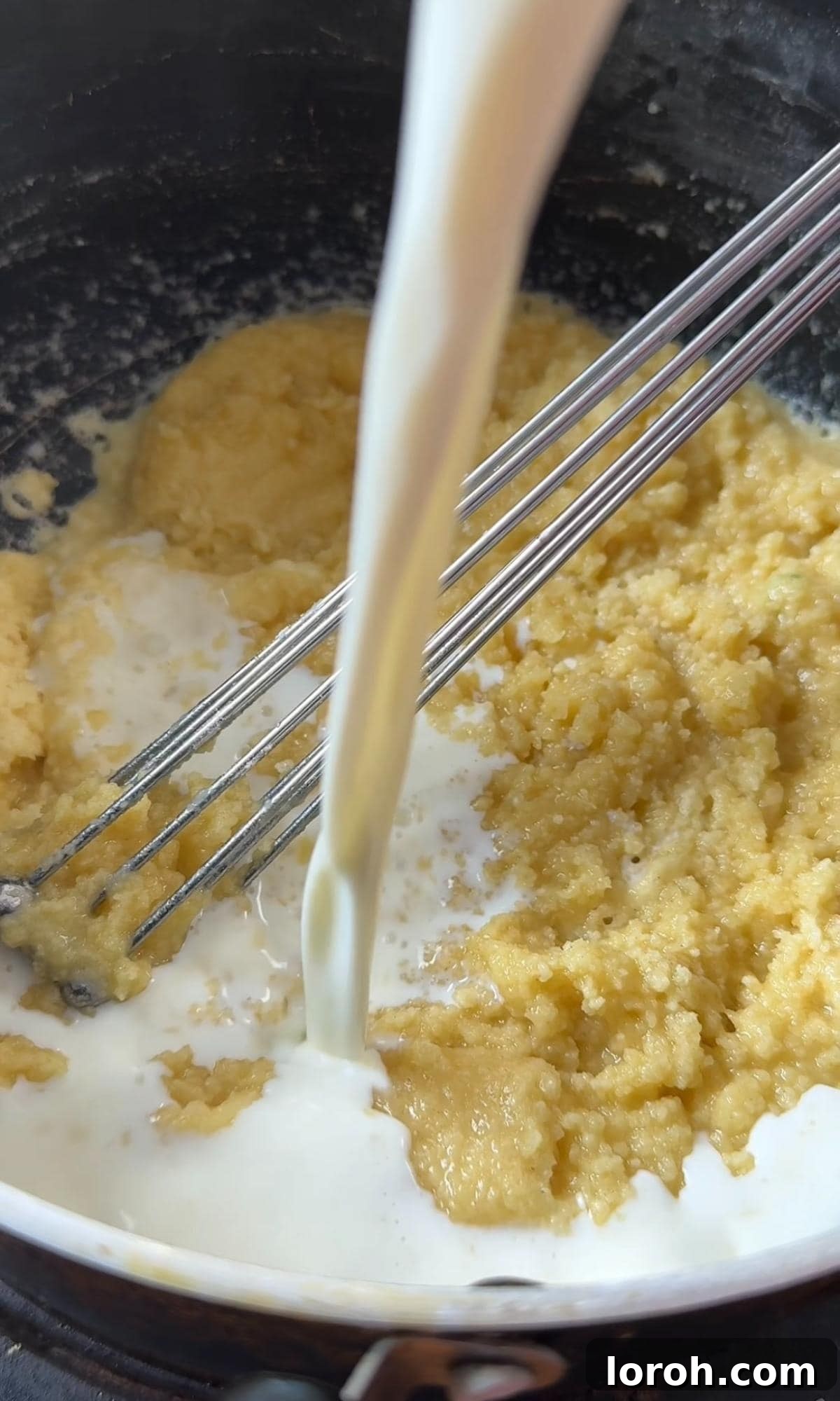 Gradually pouring milk into a mixture of melted butter and flour (roux) in a saucepan, whisking to create a smooth béchamel sauce.