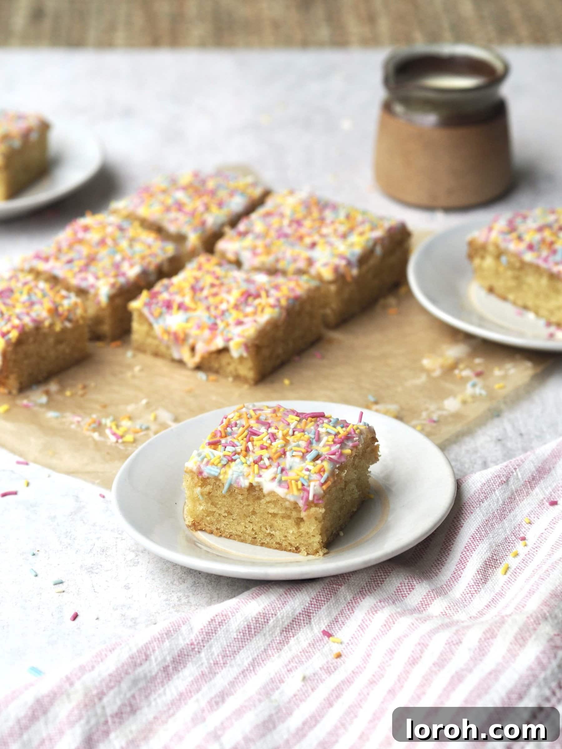 Classic Sprinkle Sponge with Golden Custard 2 A perfectly sliced piece of rainbow sprinkle-topped sponge cake on a white plate, with the rest of the beautifully baked cake in the background.