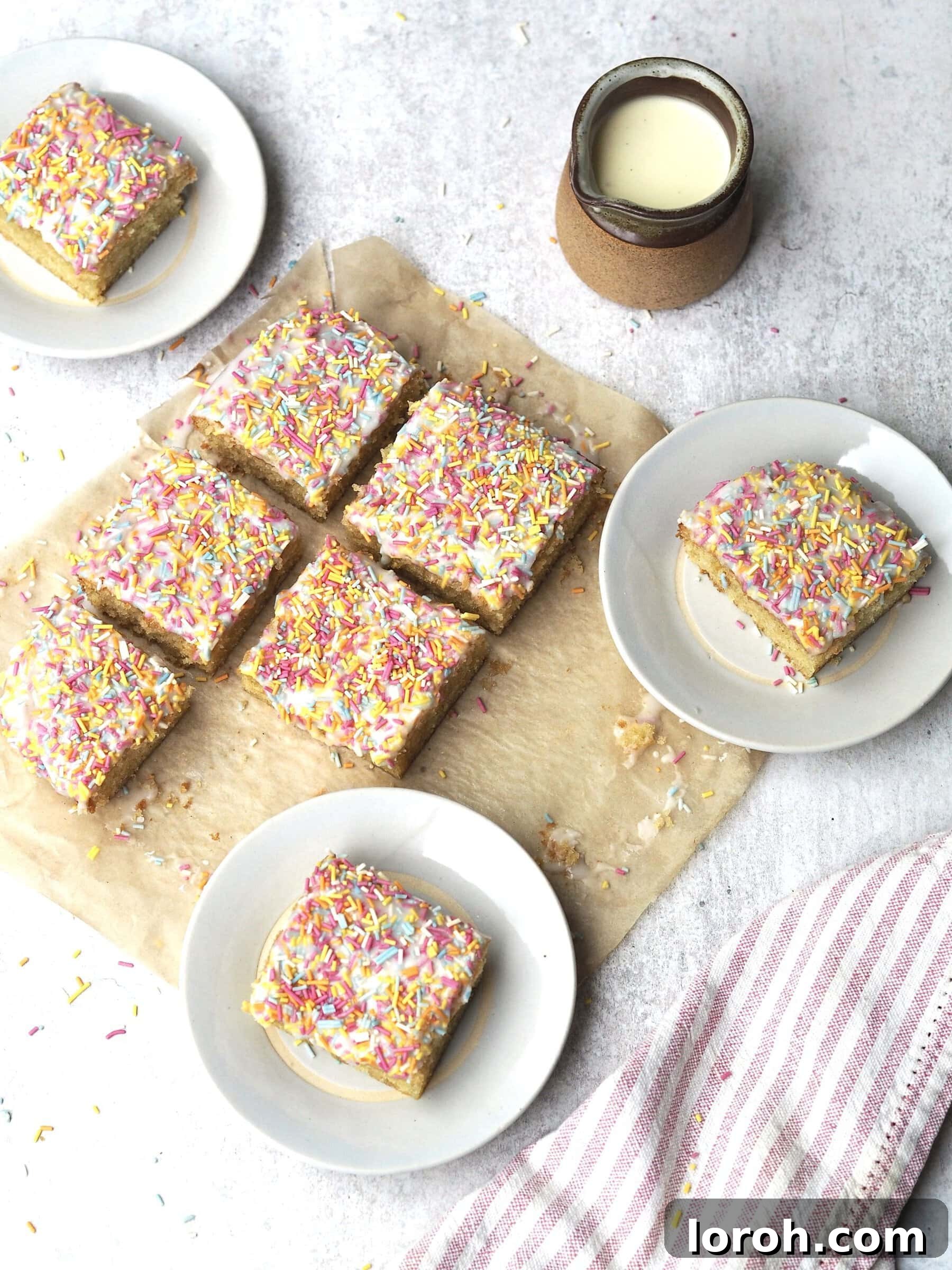 Classic Sprinkle Sponge with Golden Custard 11 Individual white plates presenting perfectly cut squares of iced sprinkle sponge cake, with the remaining cake still in the tin in the background, ready for serving.