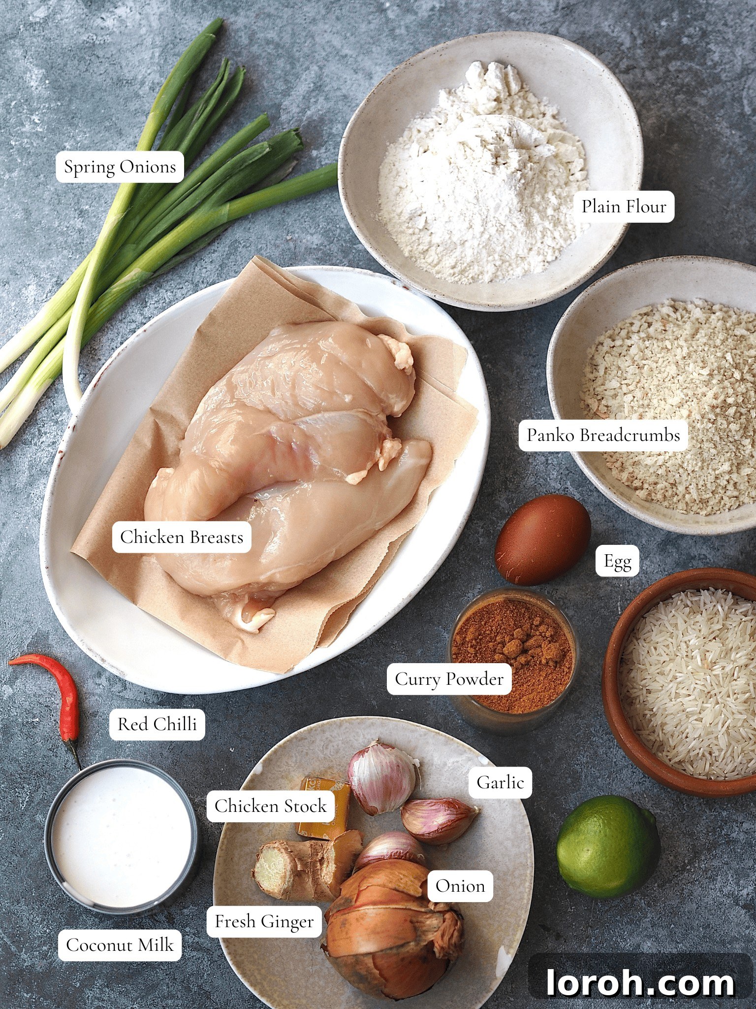 All the essential ingredients for making Chicken Katsu Curry laid out neatly on a counter, including raw chicken breasts, panko breadcrumbs, flour, eggs, sunflower oil, onion, fresh ginger, garlic cloves, curry powder, chicken stock, coconut milk, soy sauce, and a bowl of uncooked basmati rice. Fresh red chili and spring onions are also displayed for garnish.