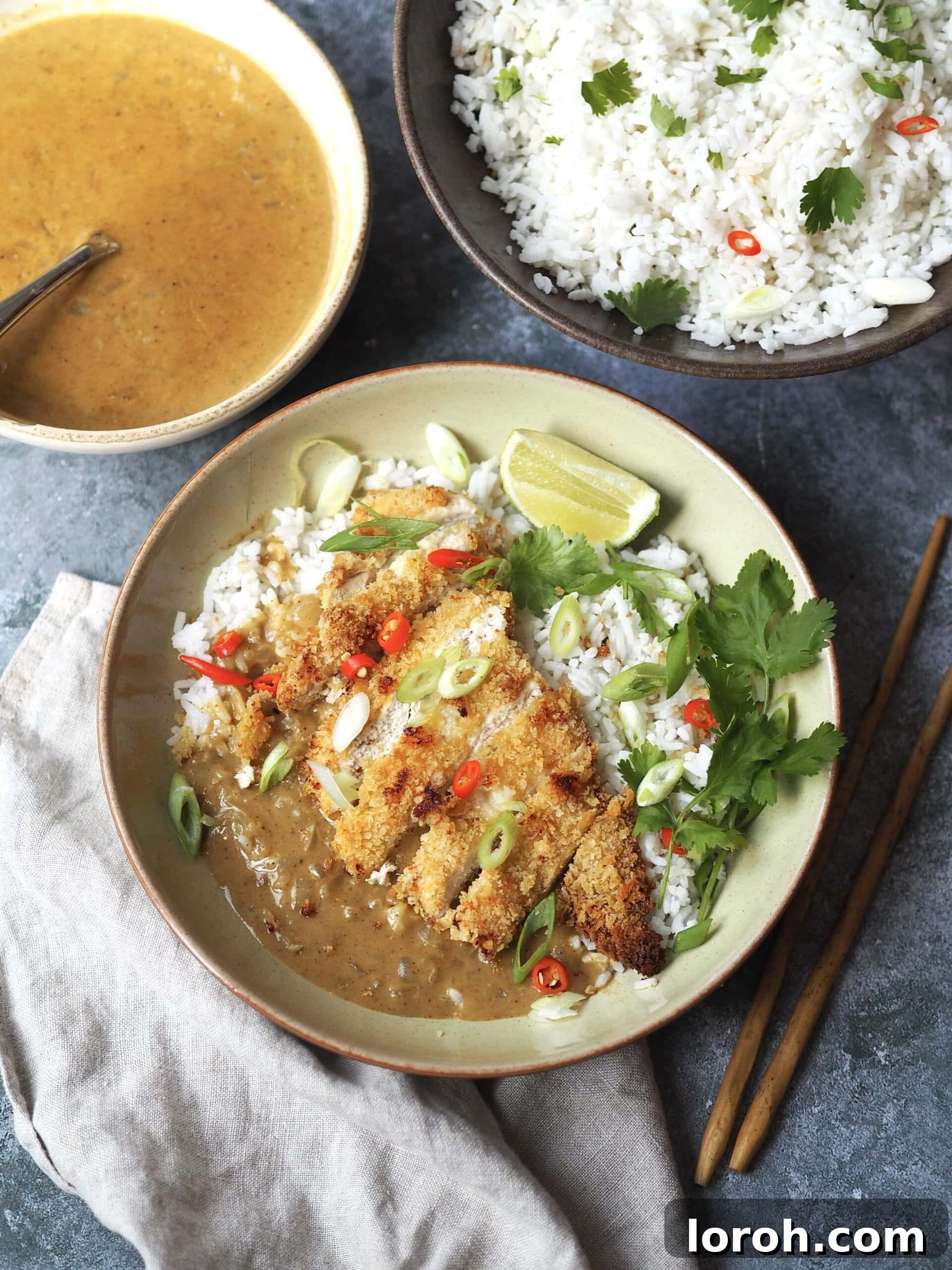 A beautifully presented chicken katsu curry bowl featuring sliced crispy chicken, aromatic rice, and a rich Japanese coconut curry sauce, artfully arranged next to separate serving bowls of herbed rice and the creamy curry sauce, ready to be enjoyed.