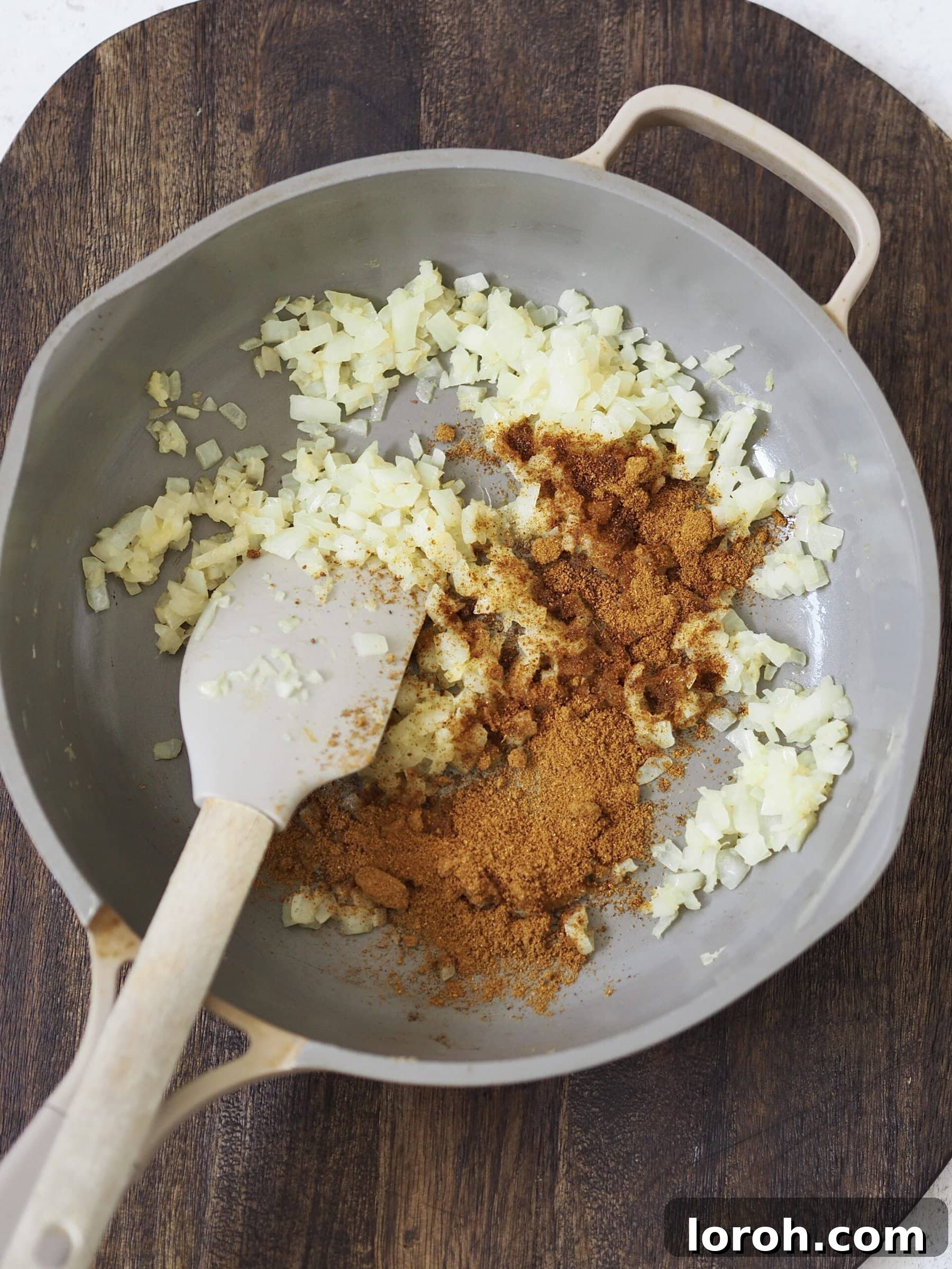 Fragrant Japanese curry powder being stirred into golden-brown fried onions, garlic, and ginger in a grey frying pan, forming the aromatic base of the curry sauce.