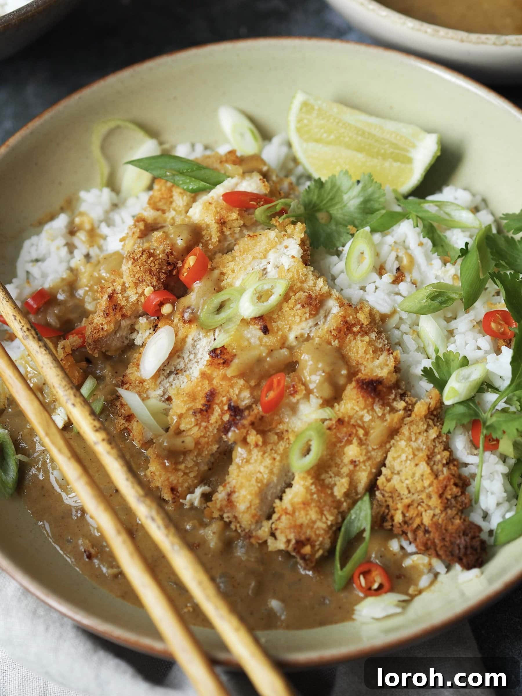 A close-up shot of a steaming chicken katsu curry rice bowl, featuring perfectly sliced crispy chicken cutlets, fluffy rice, and a generous pour of golden curry sauce, garnished with fresh spring onions and red chili. A pair of elegant wooden chopsticks rests alongside the bowl.