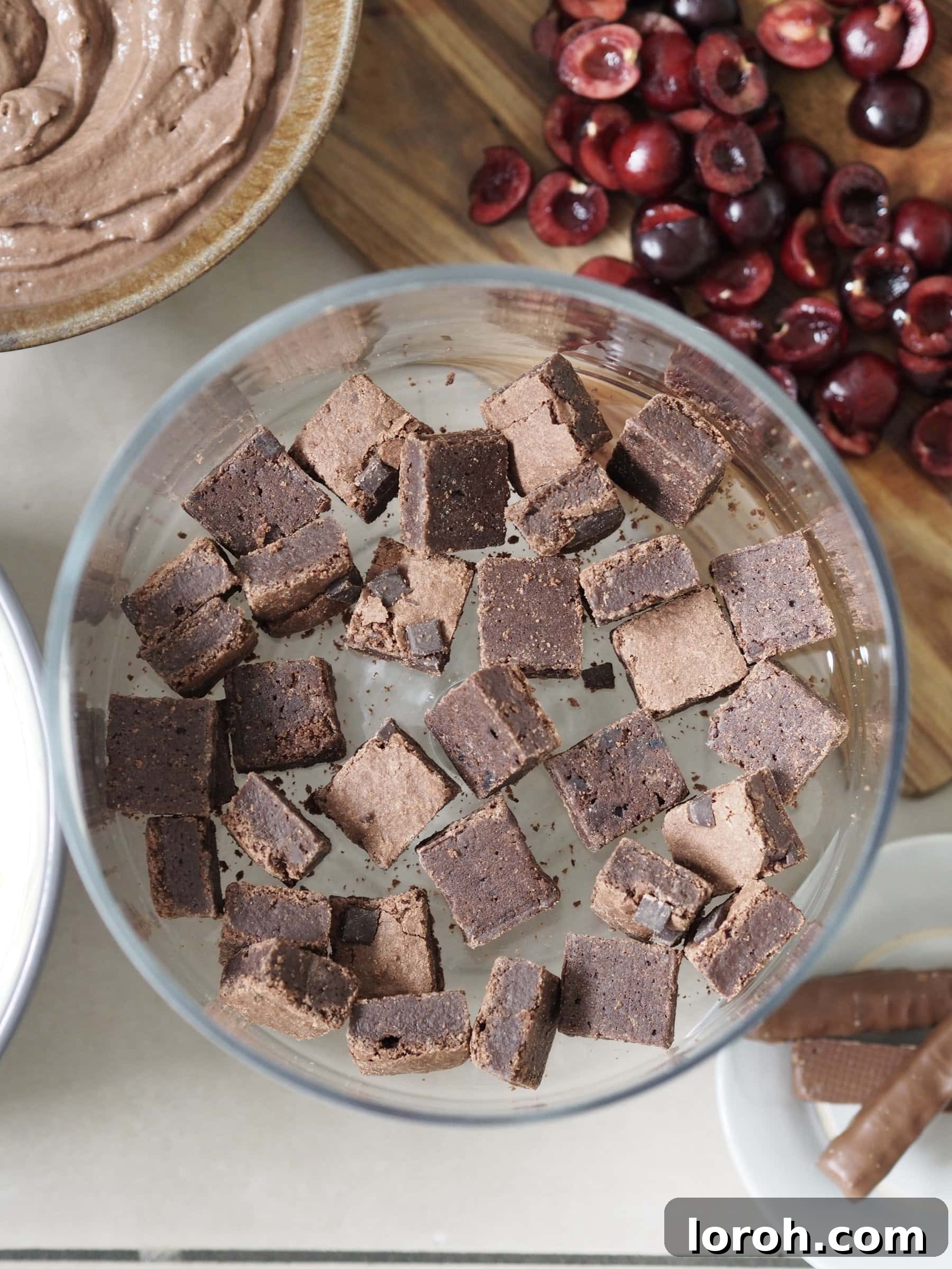 Small, fudgy brownie bites being carefully layered into the bottom of a clear glass trifle dish, forming the base.