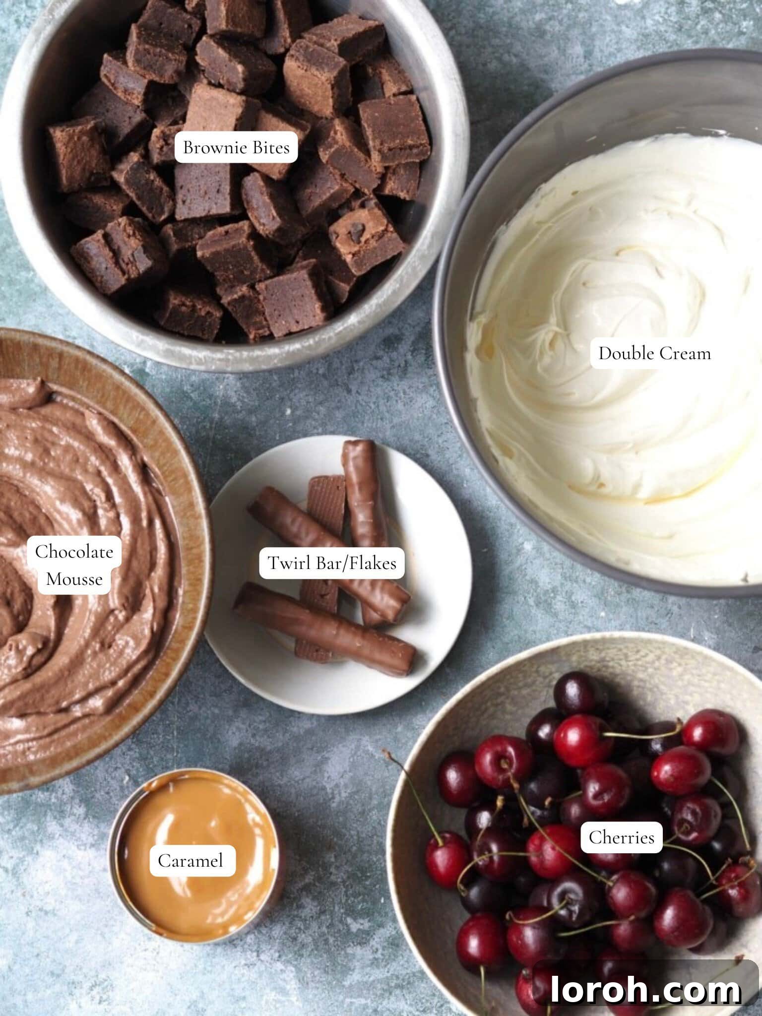 Collection of key ingredients including double cream, brownie bites, chocolate mousse, caramel, and fresh cherries, laid out for making a Chocolate Brownie Trifle.