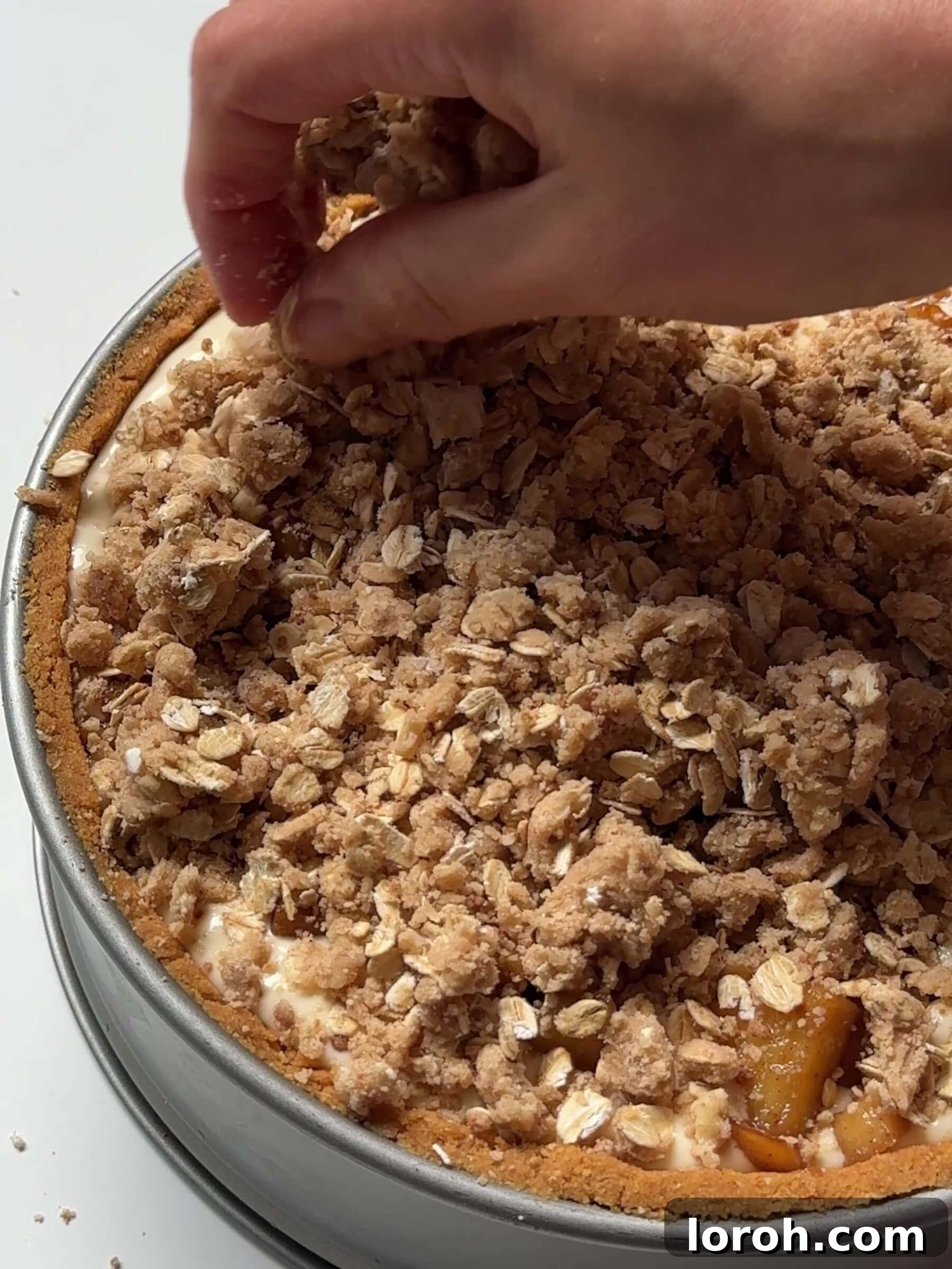 The oat crumble topping being sprinkled generously over the apple and cheesecake layers, ready for baking.
