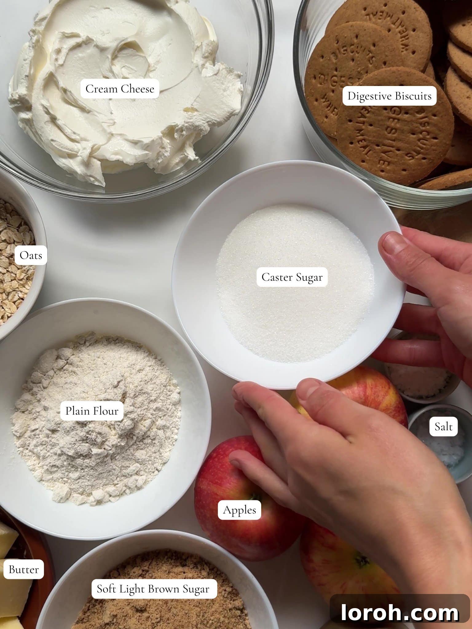 An array of fresh ingredients laid out on a table, including apples, cream cheese, butter, and biscuits, ready for making Apple Crumble Cheesecake.