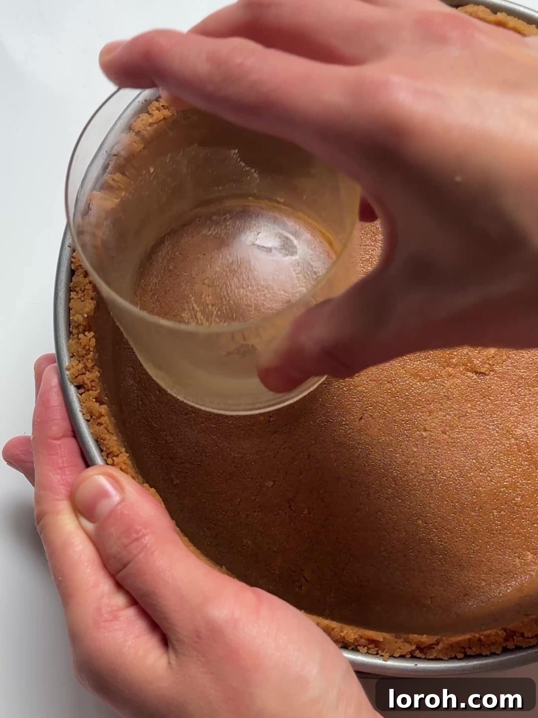 The biscuit mixture being pressed firmly into the bottom and up the sides of a baking tin using the back of a spoon.