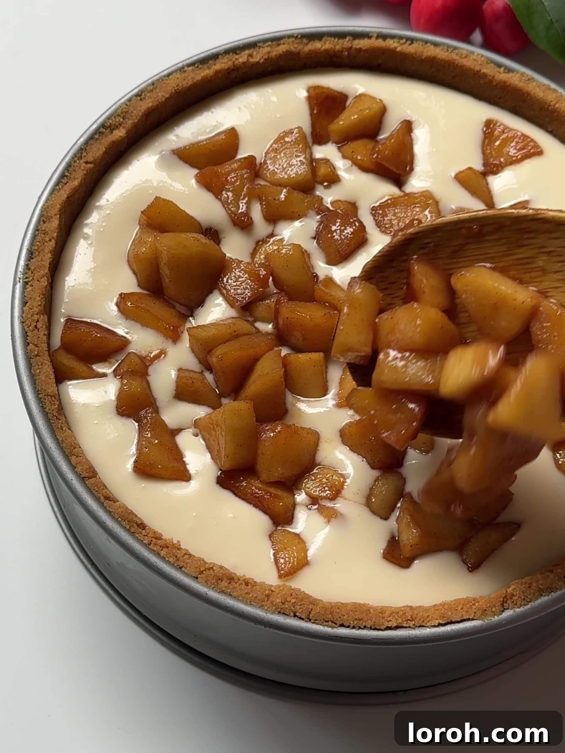 Cinnamon-spiced apples being spooned over a layer of creamy cheesecake filling in a springform tin.