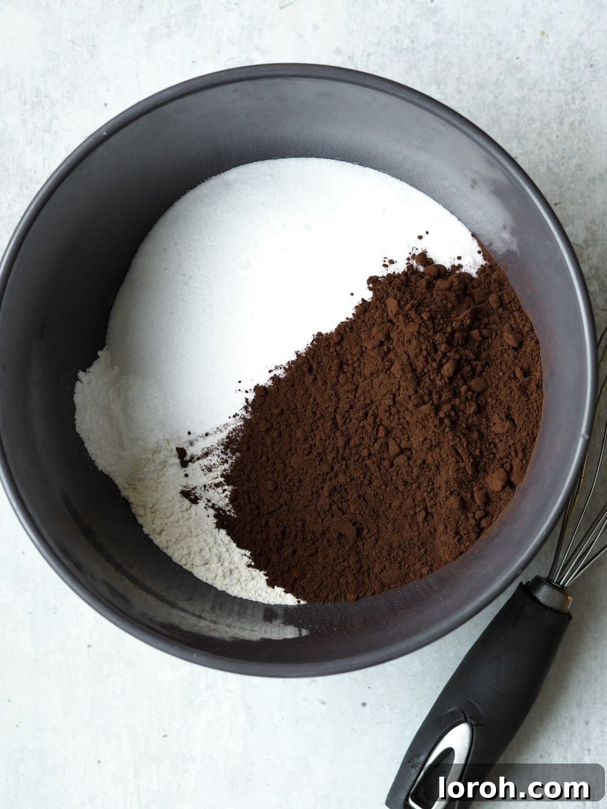 A close-up image of cocoa powder being added to caster sugar in a mixing bowl.