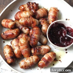 Sausages wrapped in bacon on a white serving plate next to a bowl of cranberry sauce.