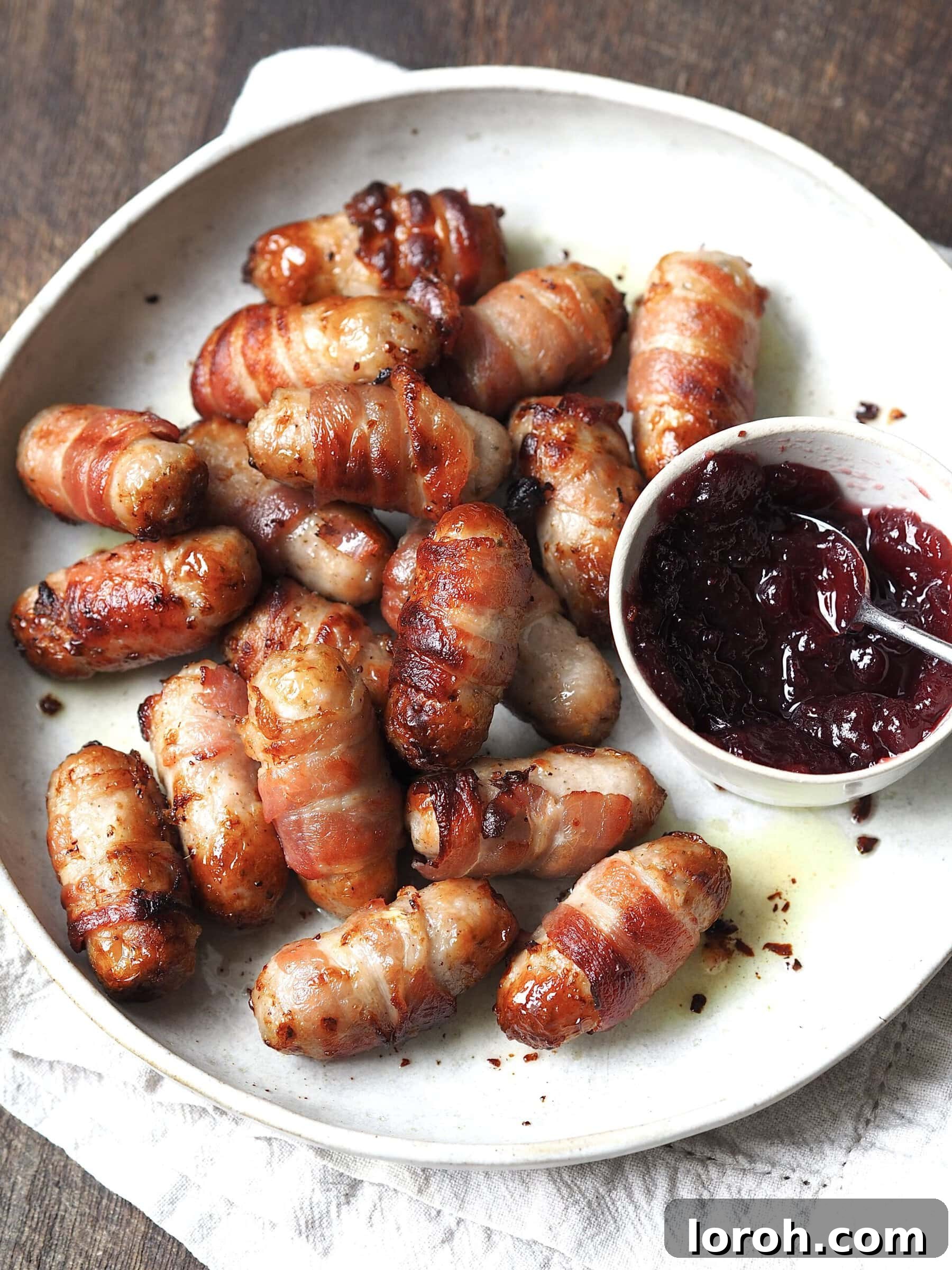 Golden Air Fryer Sausage Bites 4 Sausages wrapped in bacon on a white serving plate next to a bowl of cranberry sauce.