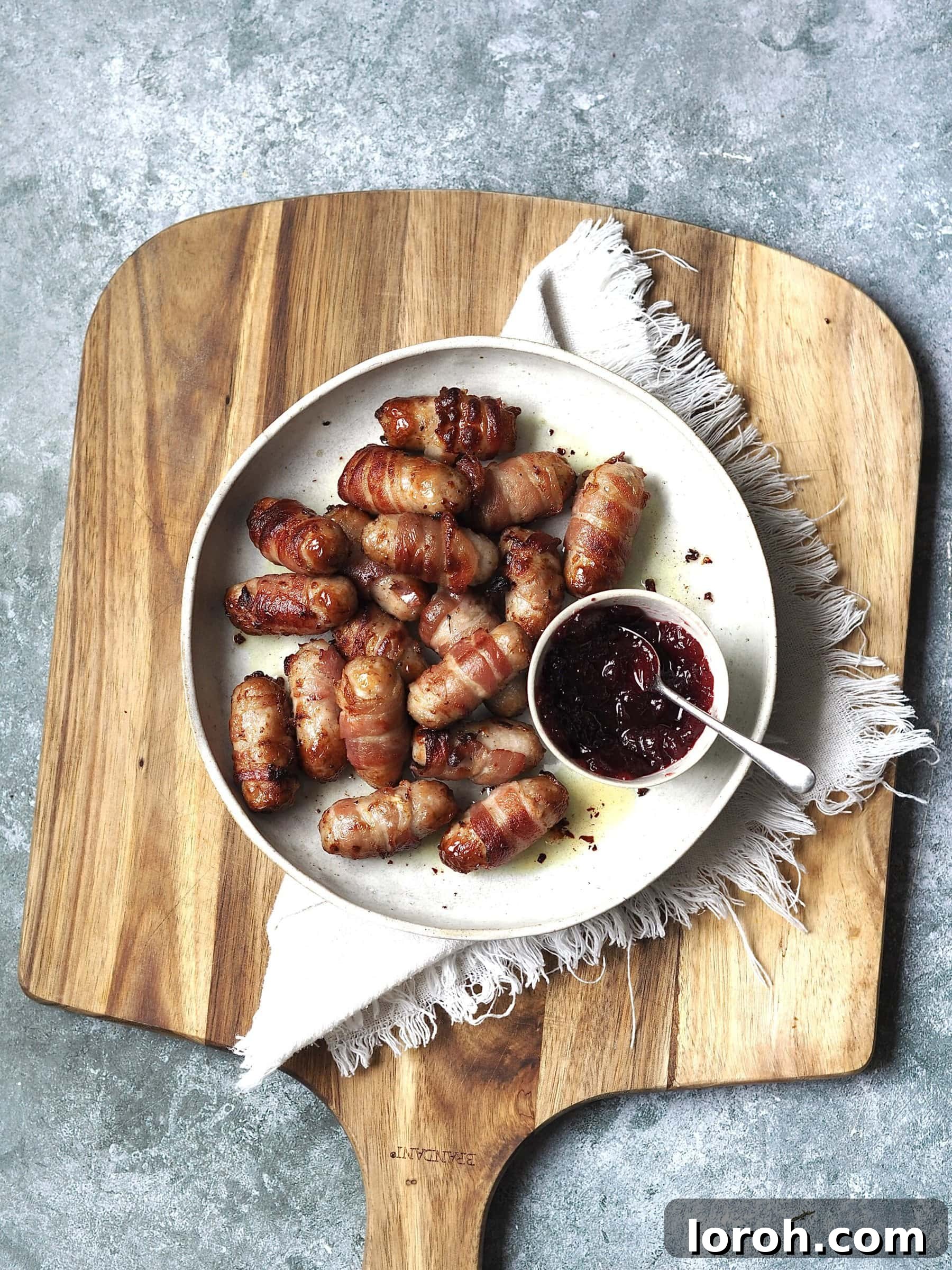 Golden Air Fryer Sausage Bites 7 Wooden board with a plate of sausages wrapped in bacon with a dipping bowl of cranberry sauce.
