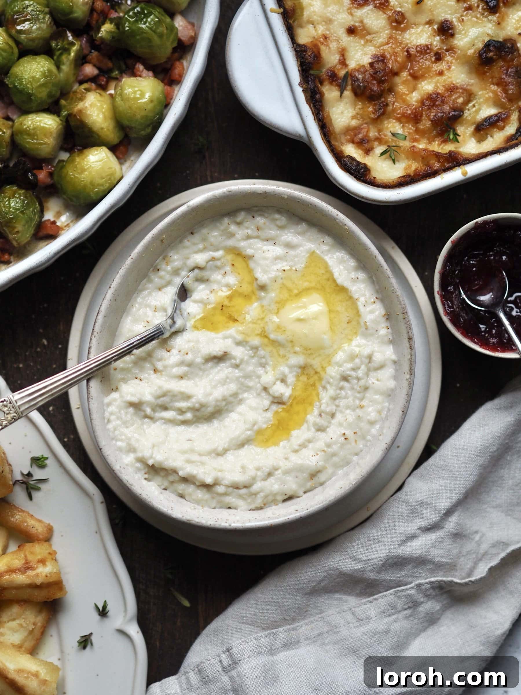 White bowl of bread sauce on a table with dishes of brussels sprouts, cheesy leeks, glazed parsnips and cranberry sauce.