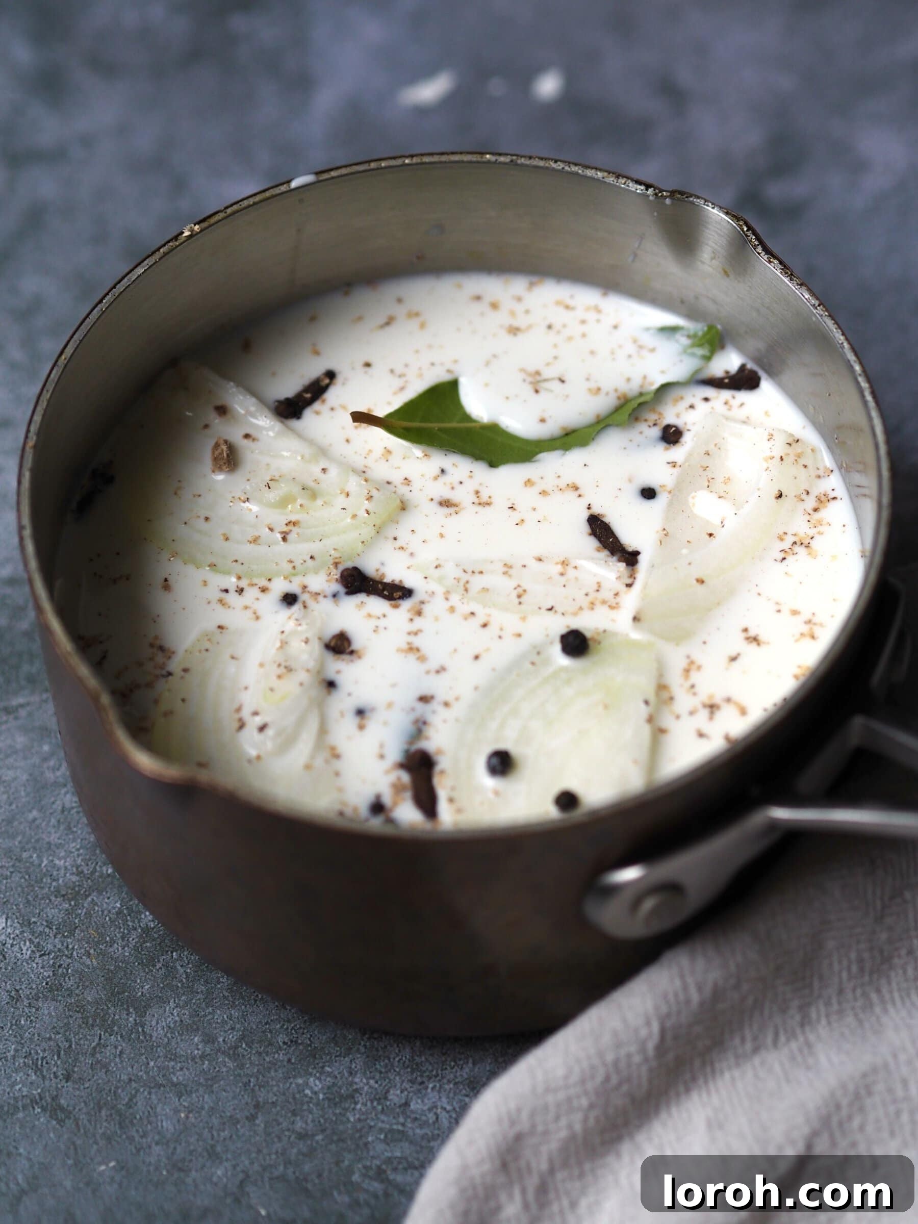 Saucepan of milk being infused with onion, peppercorns, nutmeg and a fresh bay leaf.