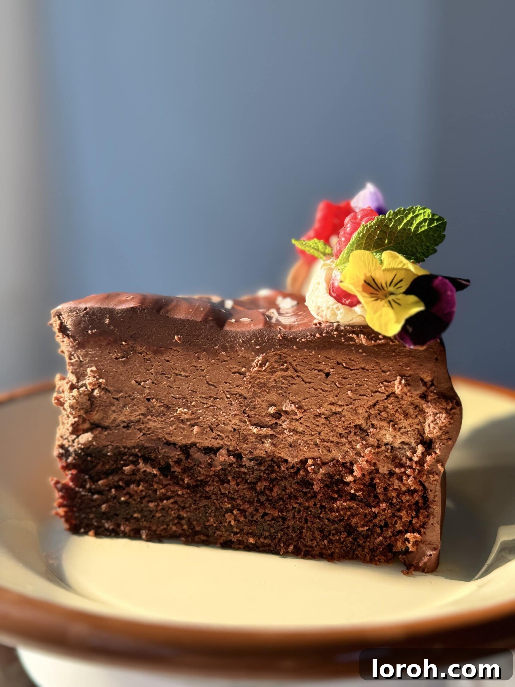 A slice of chocolate cake with chocolate mousse on a white plate, adorned with berries and edible flowers.
