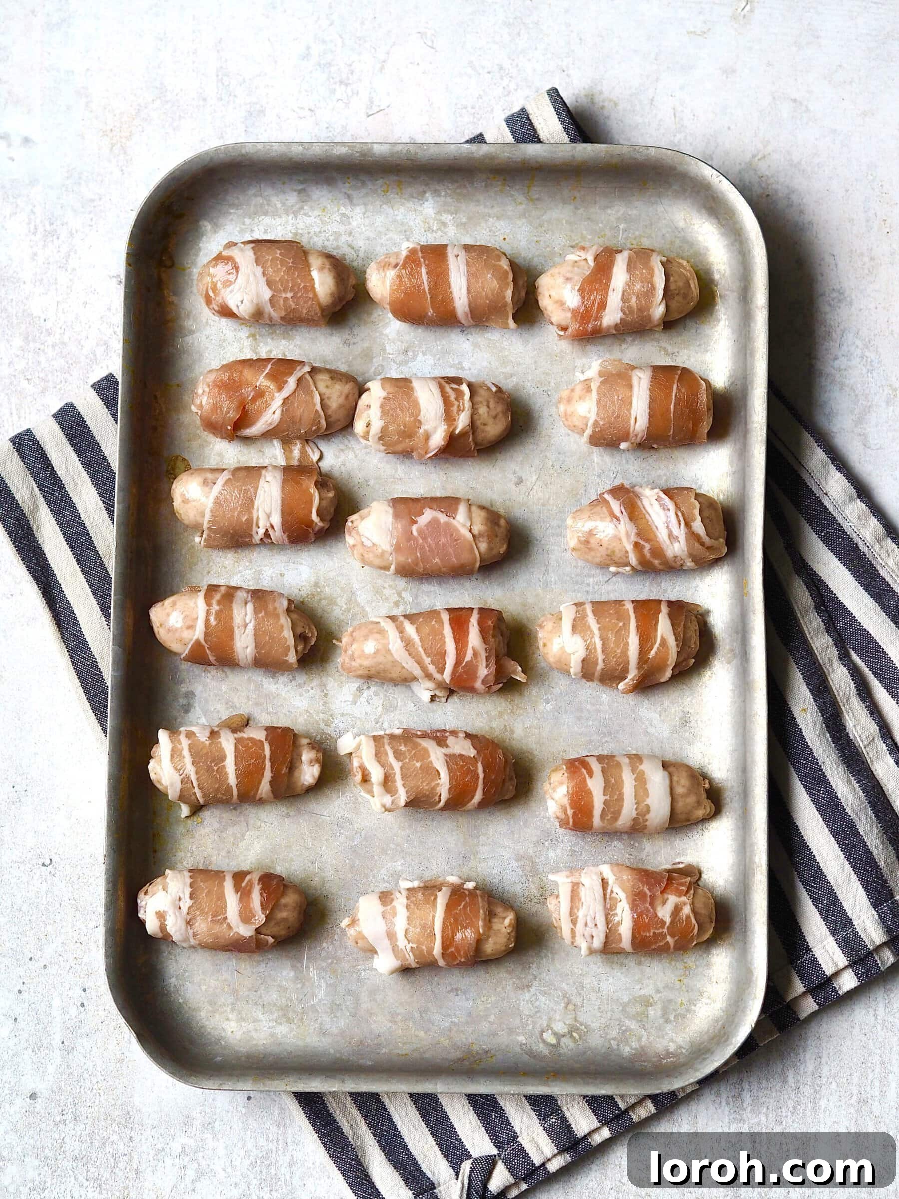 Metal roasting tray of uncooked pigs in blankets.