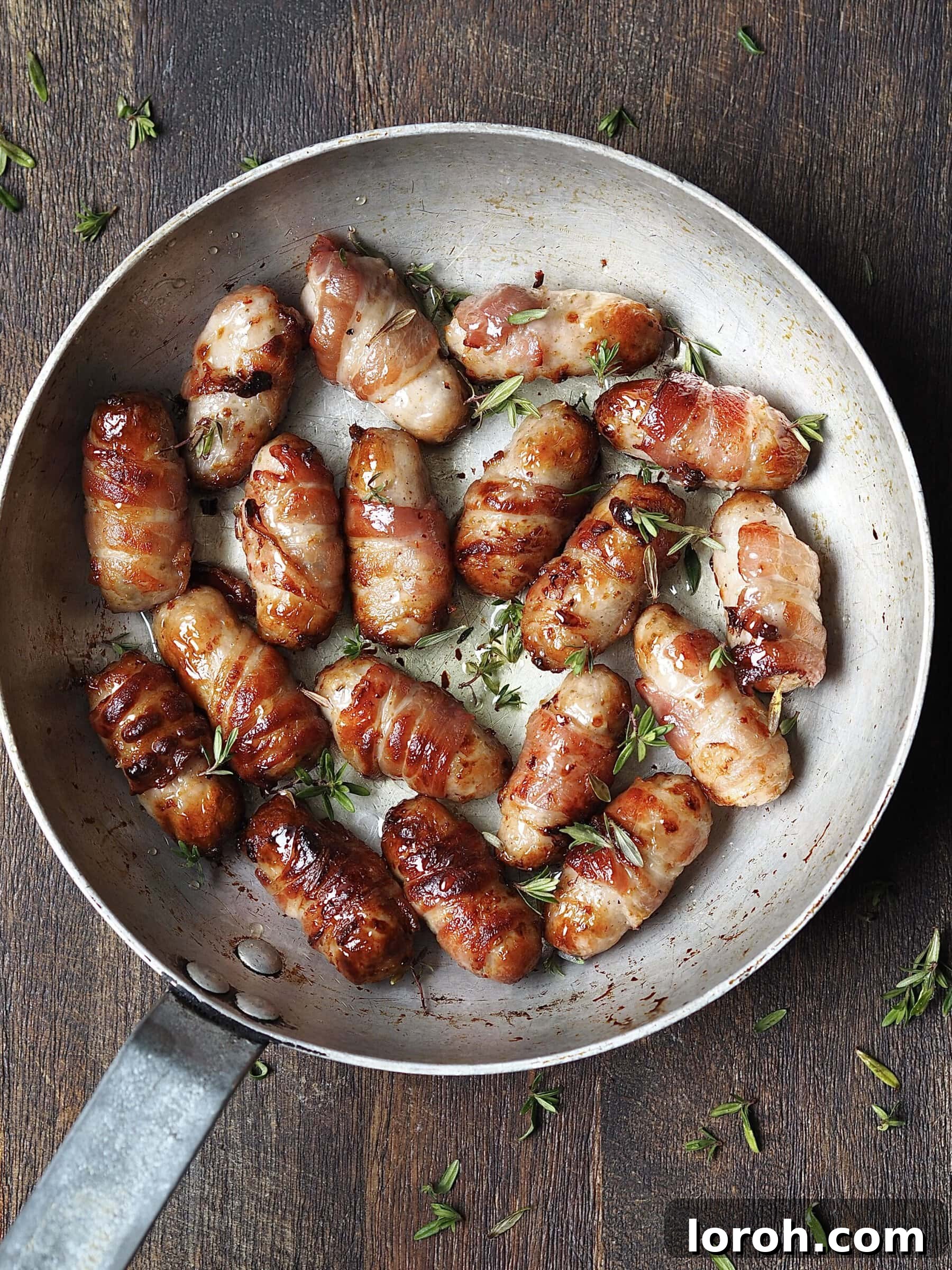 Metal saucepan on a wooden countertop with cooked pigs in blankets with a sticky fresh thyme glaze. 