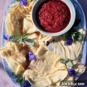 A blue plate holds beautifully folded Pannekaker (Norwegian pancakes), garnished with a vibrant raspberry compote.