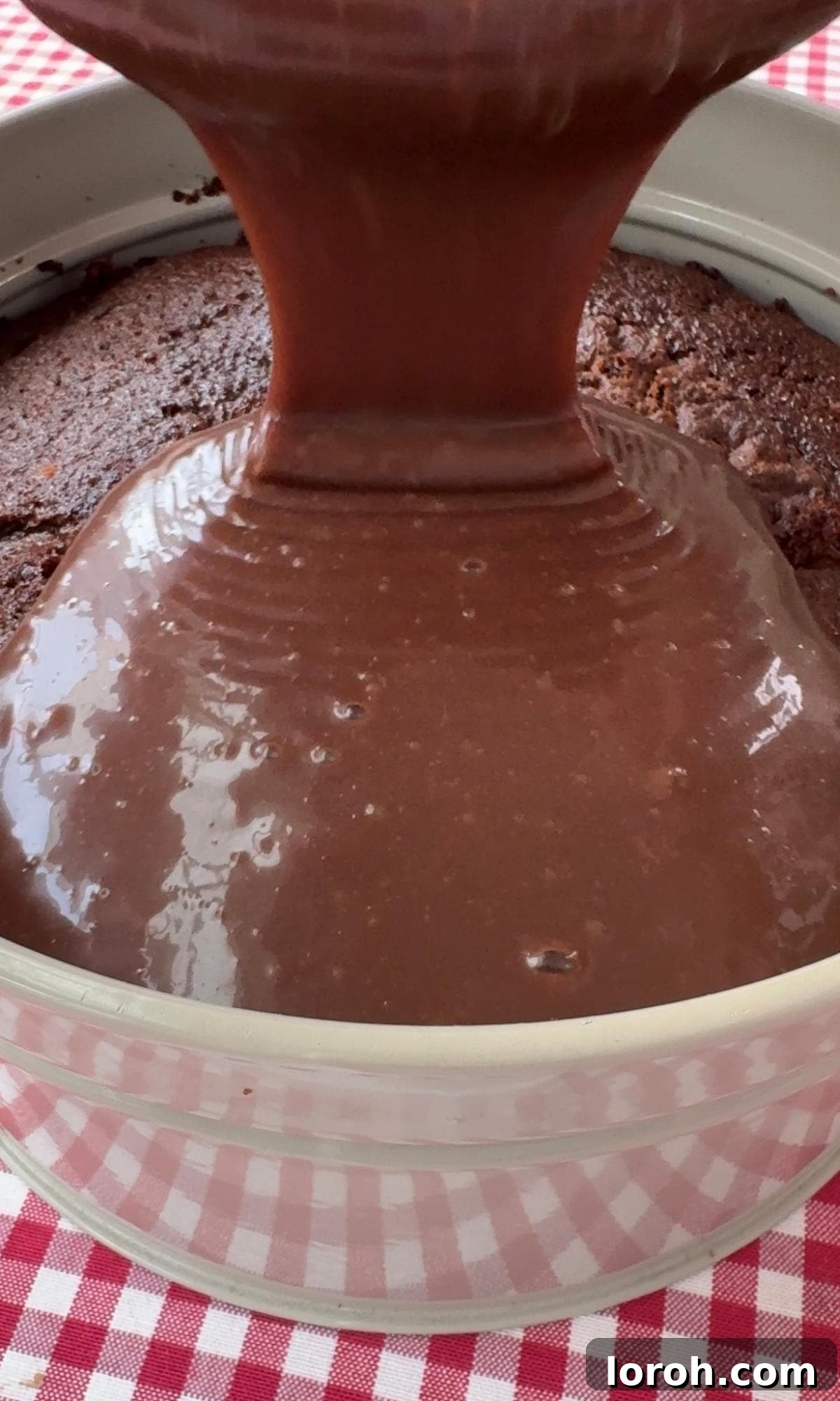 Pouring smooth chocolate ganache evenly over the completely cooled chocolate cake in its tin.
