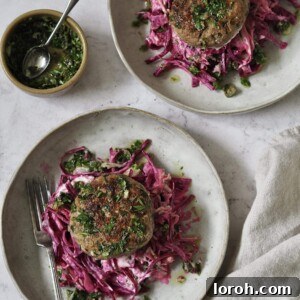 Chicken burgers on piles of pink yoghurt and beetroot slaw on grey stone plates next to a small bowl of salsa verde that's been spooned over the top.