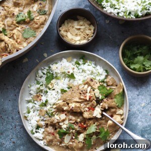 Plate of almond butter chicken curry and rice surrounded by serving dishes of rice, curry and toppings.