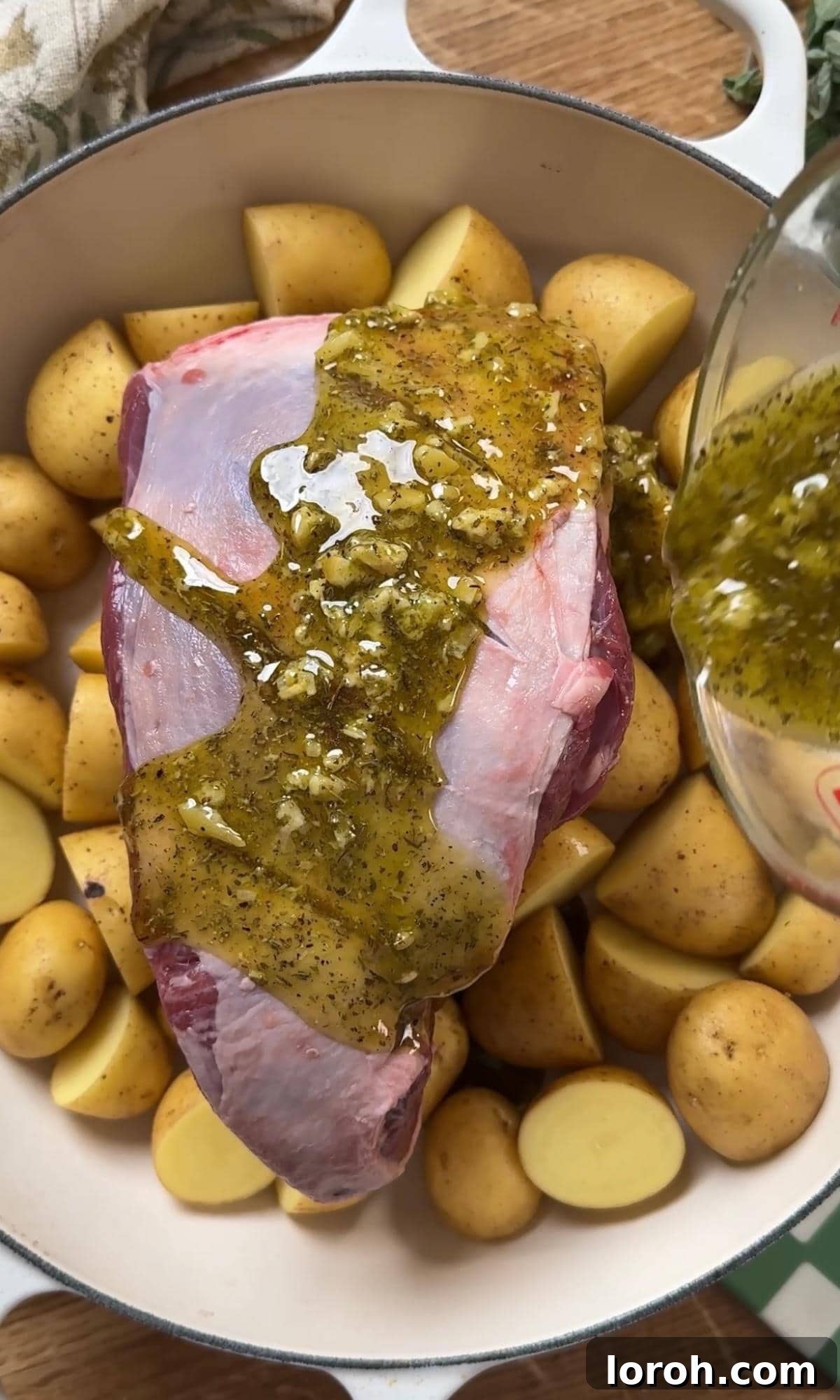 Pouring the prepared marinade over a half leg of lamb resting on baby potatoes in a Dutch oven.